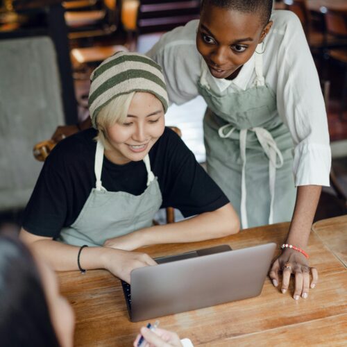 From above of optimistic diverse female workers of cafeteria discussing business plan while using laptop and taking notes in piece of paper at table